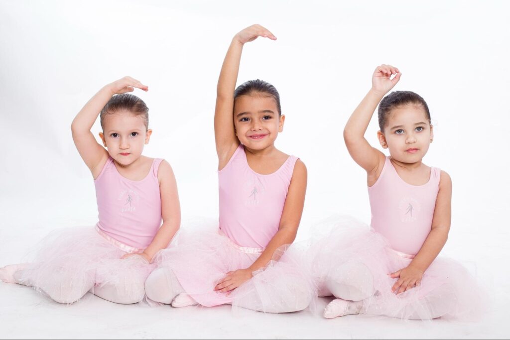 Três meninas com roupa de ballet cor de rosa, em posição de dança, sentadas no chão, olhando para a câmera