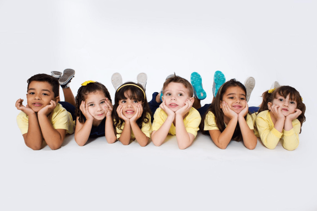 Grupo com seis alunos da educação infantil uniformizados, deitados no chão, segurando o queixo e posando para a foto adquirindo competências socioemocionais