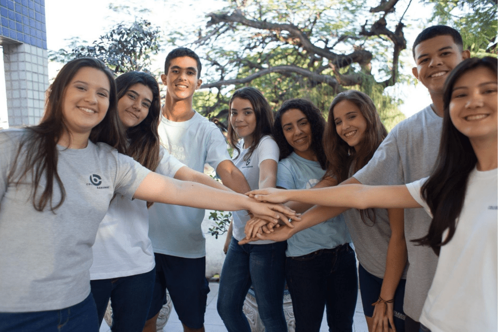 Grupo com alunos do ensino médio, ao ar livre, fazendo sinal de união com as mãos uma sobre a outra.