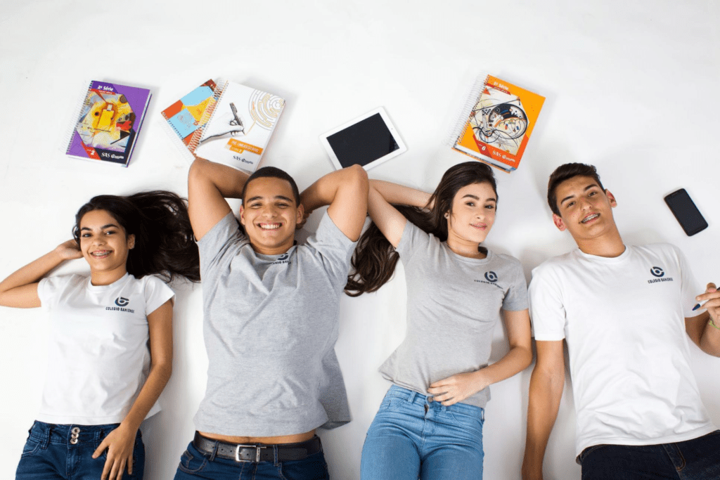 Grupo com 4 jovens do ensino médio deitados no chão, sorrindo junto a livros.