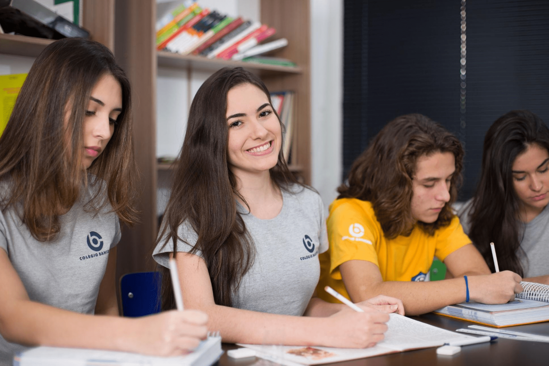 Grupo com quatro alunos sentados e uniformizados, sendo apenas uma aluna central em foco e sorrindo para a câmera