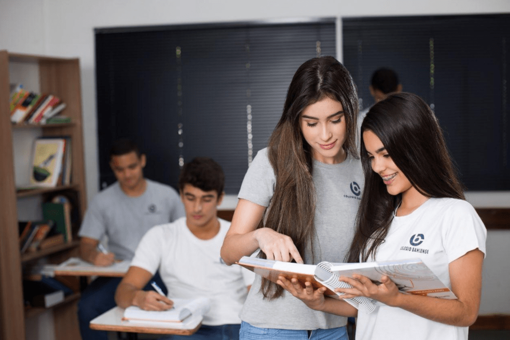 Grupo de estudantes em sala de aula, sendo dois sentados escrevendo ao fundo e mais duas alunas em foco olhando para o conteúdo do livro em mãos