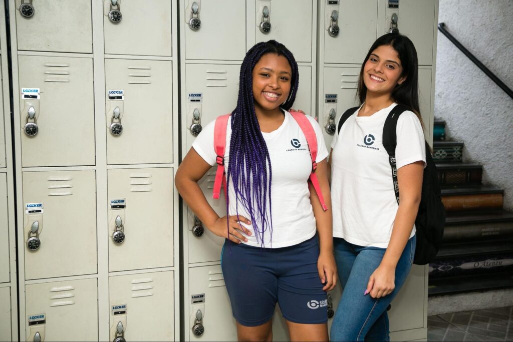 Alunas do Ensino Médio, em frente aos armários, sorrindo para a câmera