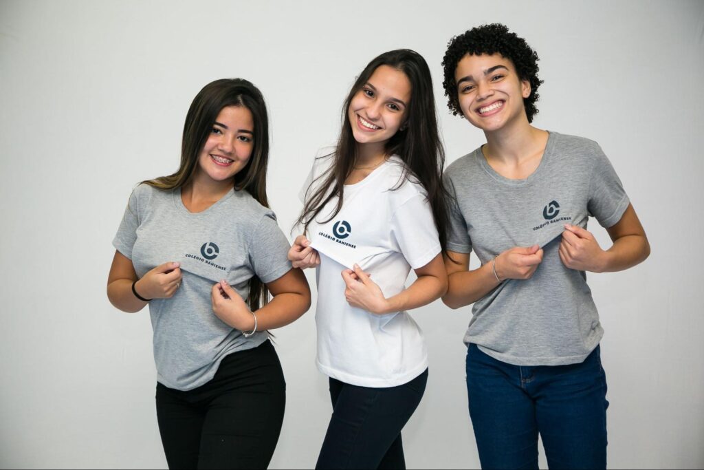 Três alunas do Ensino Médio do Colégio Bahiense sorrindo para câmera enquanto seguram o uniforme escolar evidenciando a logo do Bahiense