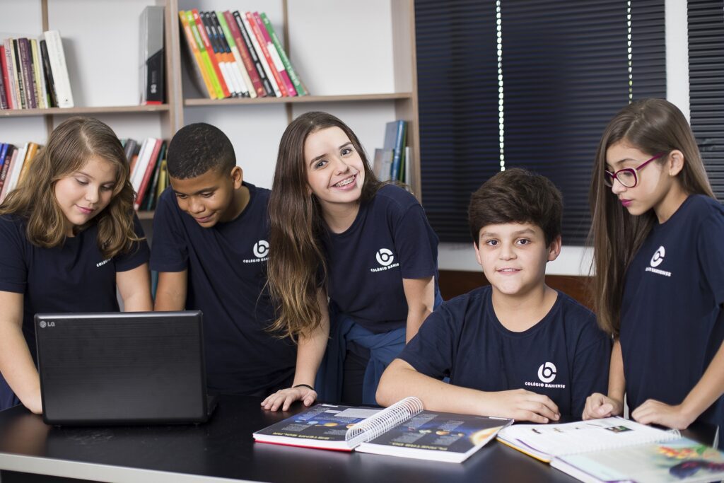 Grupo com 5 alunos uniformizados diante de livros e de um computador