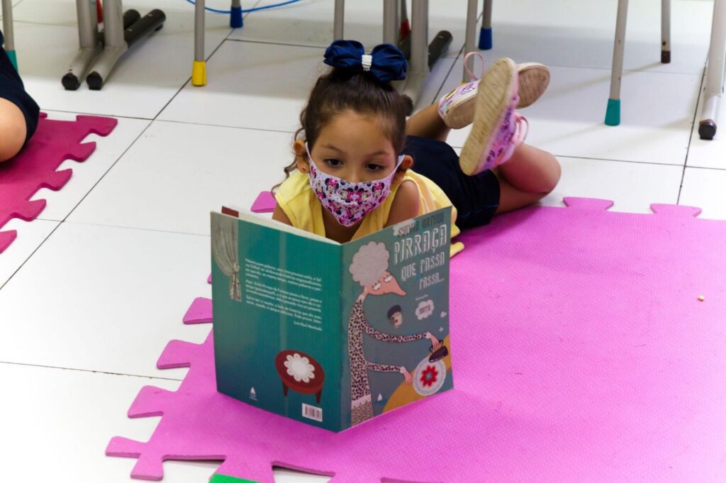 livros para crianças: menina de máscara lendo livro infantil