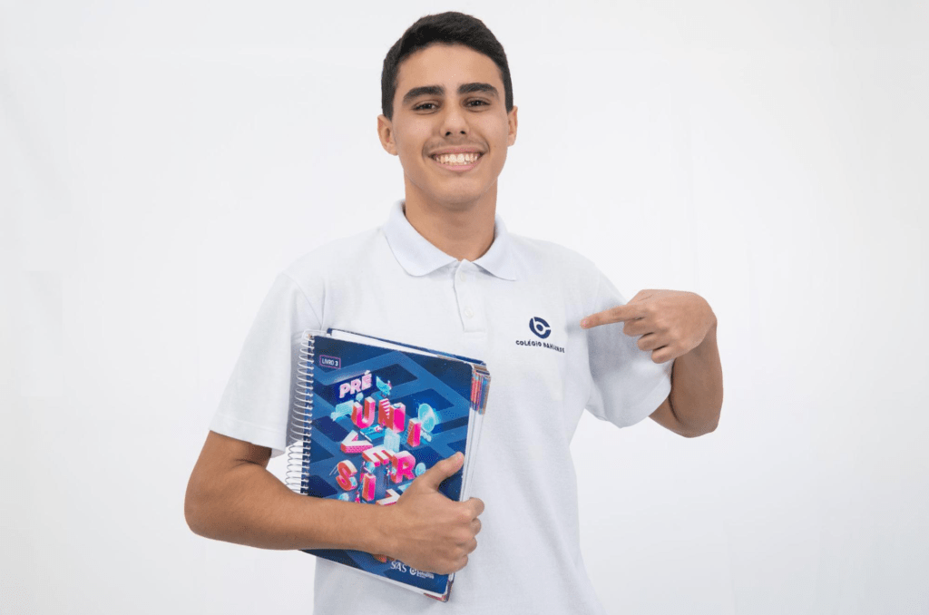 Aluno do Colégio Bahiense segurando caderno e posando para foto enquanto sorri e aponta para a logo do Colégio Bahiense em sua camisa branca de uniforme